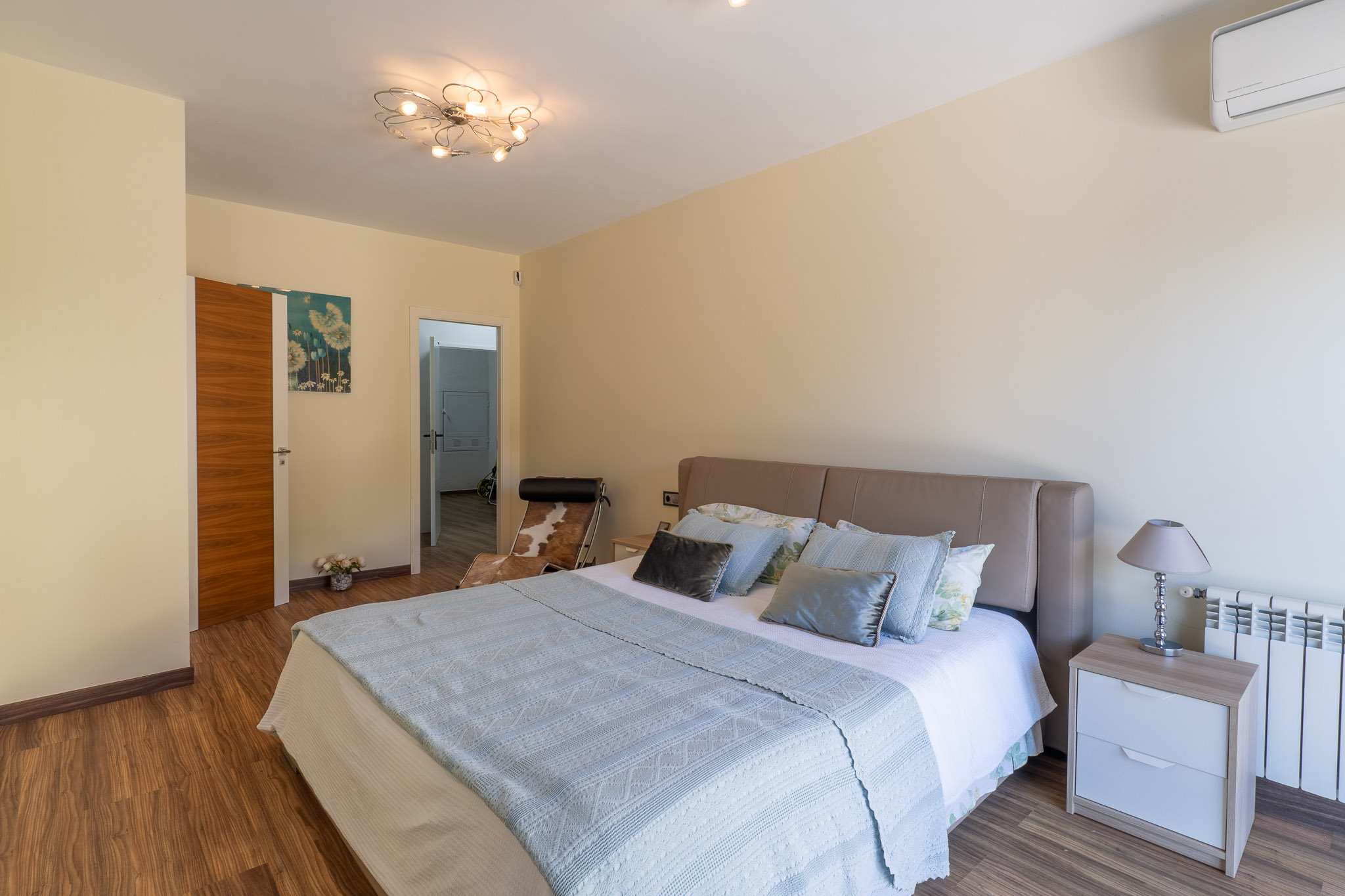 Dormitorio con cama de cabecero beige, suelo laminado de madera y moderna lámpara de techo. Incluye mesita de noche con lámpara, radiador y aire acondicionado. Dormitorio con cama de cabecero beige, suelo laminado de madera y moderna lámpara de techo. Incluye mesita de noche con lámpara, radiador y aire acondicionado.