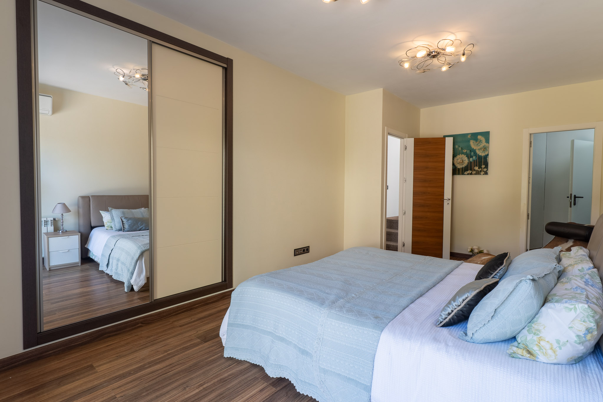 Dormitorio con cama de textiles azules, suelo laminado de madera y armario corredero con panel espejado. Destaca la moderna lámpara de techo y un cuadro floral. Dormitorio con cama de textiles azules, suelo laminado de madera y armario corredero con panel espejado. Destaca la moderna lámpara de techo y un cuadro floral.