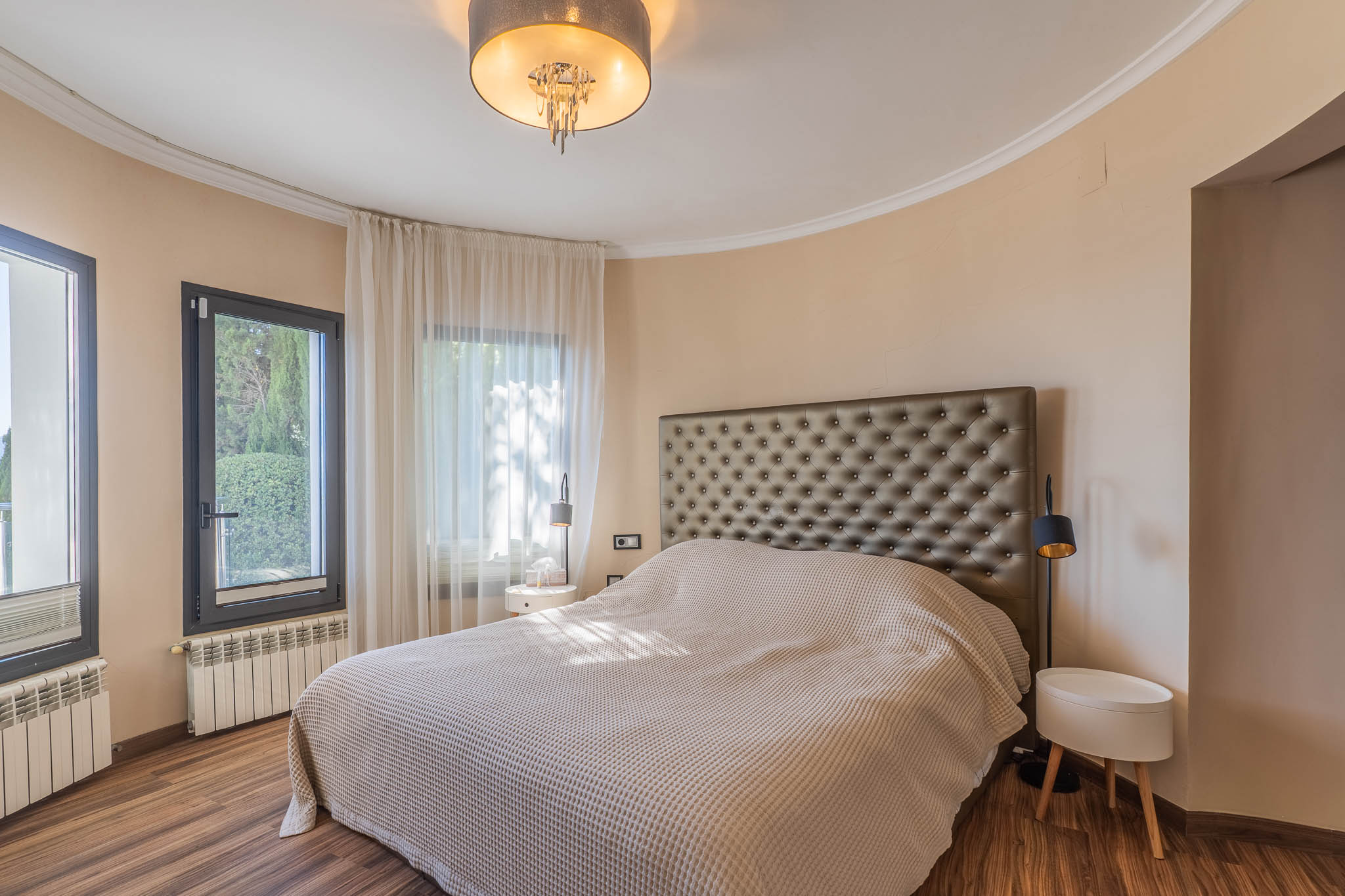 Dormitorio con cama grande y cabecero tapizado. Pared curva con ventanales de marco oscuro y cortinas claras. Suelo de madera, radiadores y lámpara de techo moderna. Dormitorio con cama grande y cabecero tapizado. Pared curva con ventanales de marco oscuro y cortinas claras. Suelo de madera, radiadores y lámpara de techo moderna.