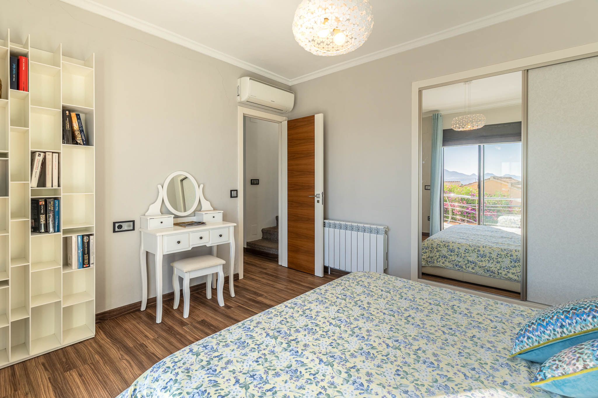 Dormitorio con suelo laminado de madera, tocador blanco y estantería modular. El armario con espejo refleja una vista a la montaña desde el balcón. Dormitorio con suelo laminado de madera, tocador blanco y estantería modular. El armario con espejo refleja una vista a la montaña desde el balcón.