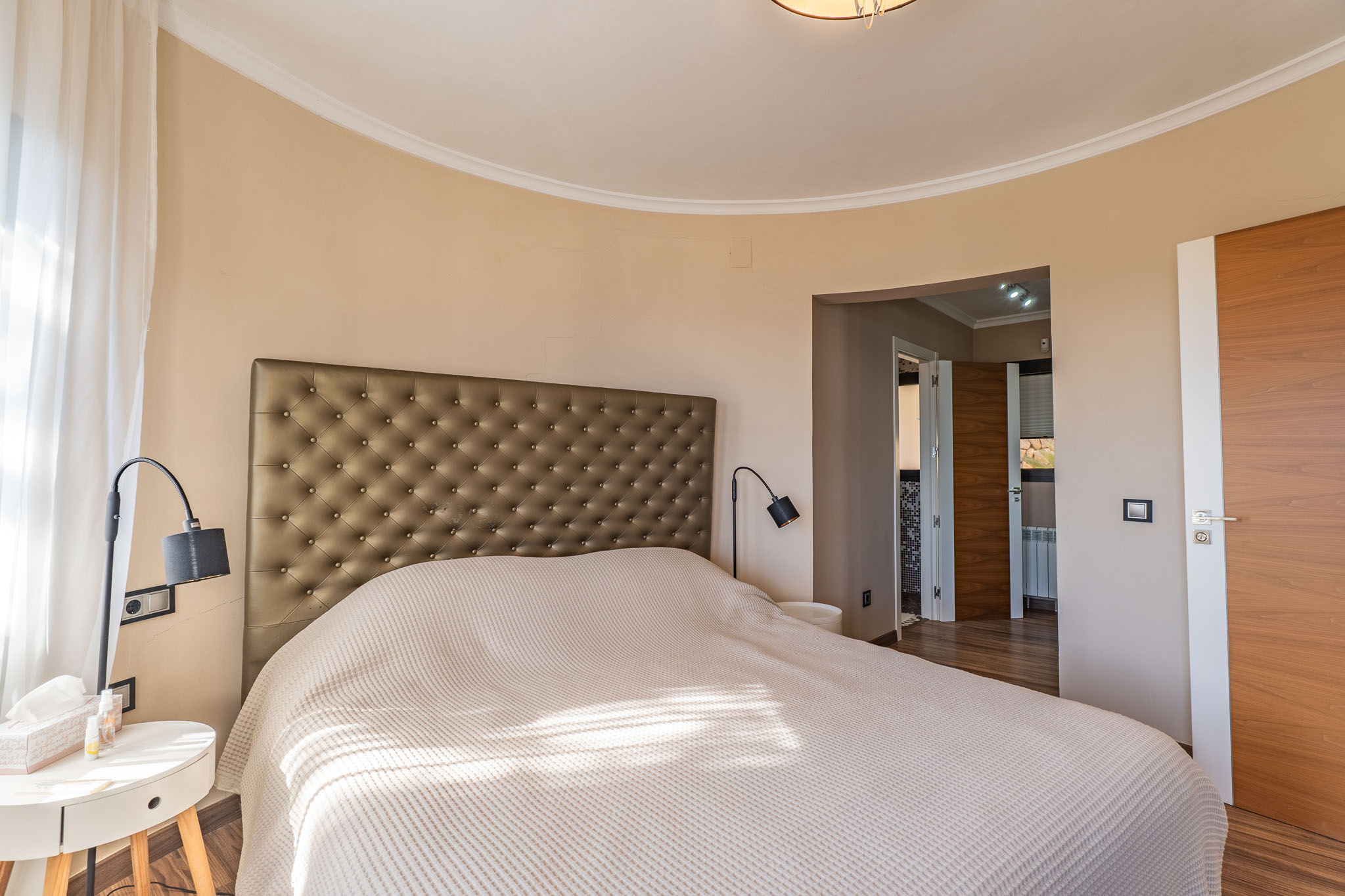 Dormitorio principal con cabecero tapizado, pared curva y luz natural filtrada por cortinas translúcidas, complementado con puertas de madera. Dormitorio principal con cabecero tapizado, pared curva y luz natural filtrada por cortinas translúcidas, complementado con puertas de madera.