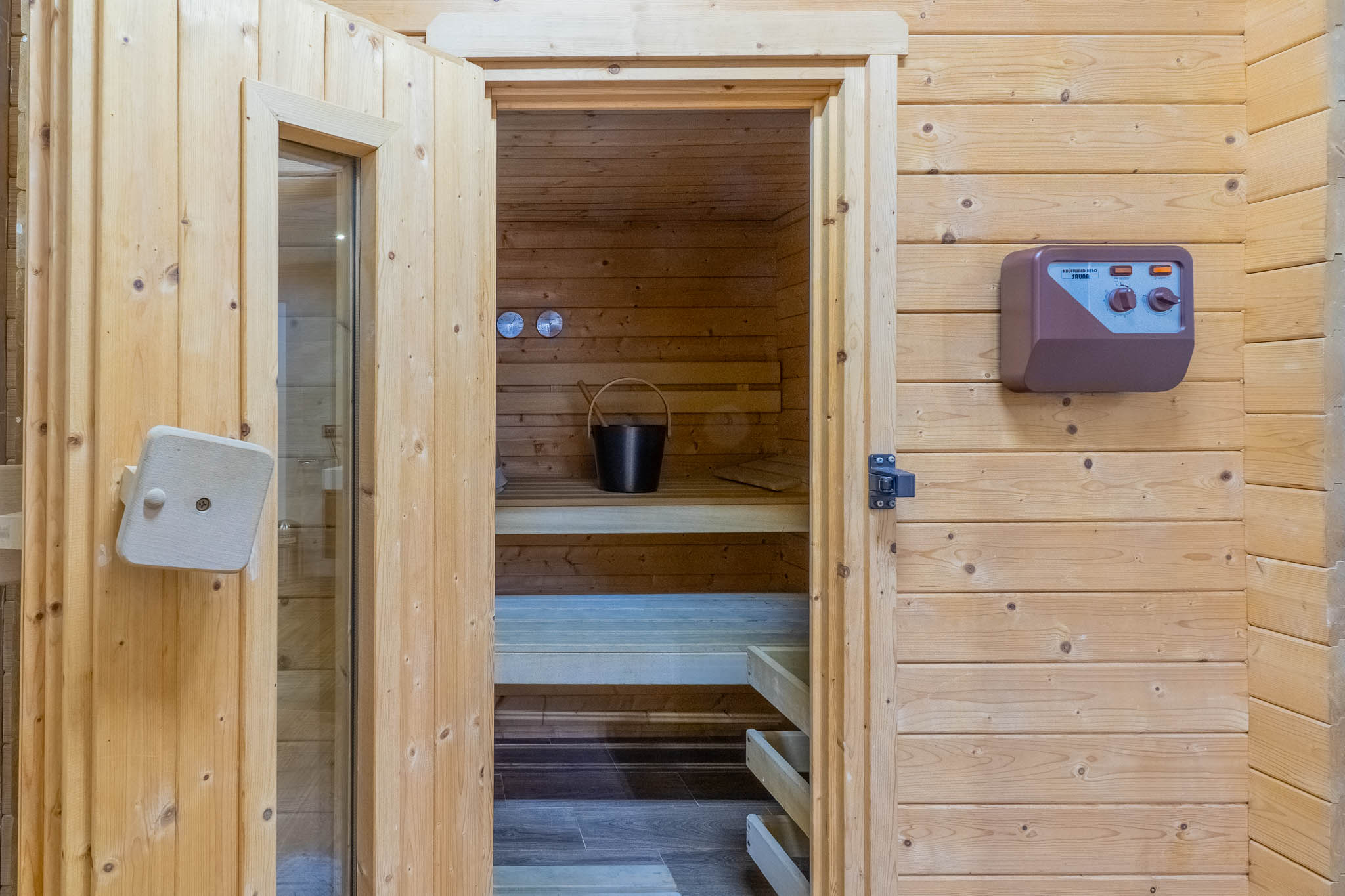 Entrada a la sauna revestida en madera clara, mostrando bancos interiores, un cubo con cucharón y un panel de control externo. Entrada a la sauna revestida en madera clara, mostrando bancos interiores, un cubo con cucharón y un panel de control externo.
