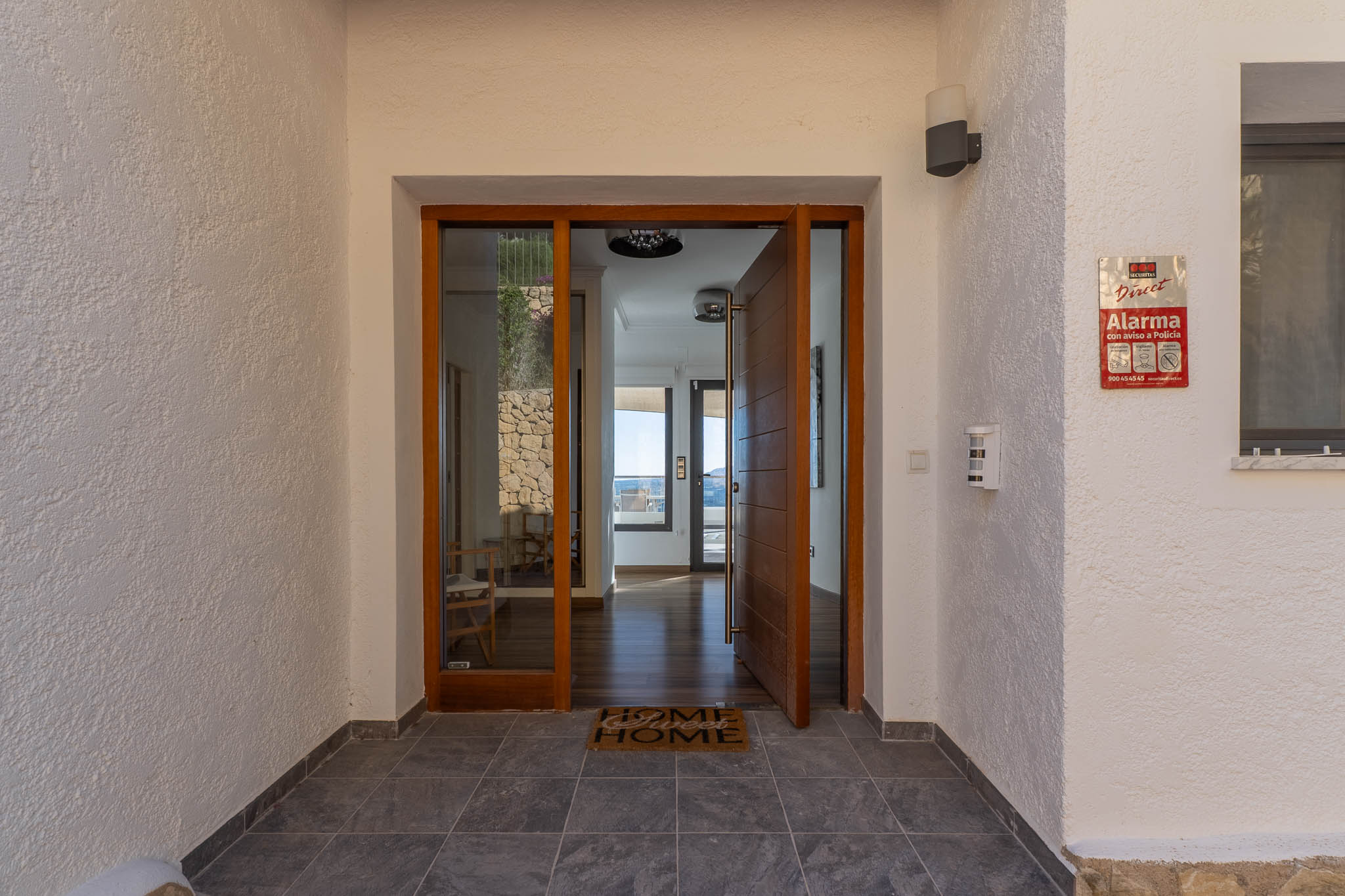 Entrada principal con puerta pivotante de madera y panel de cristal, revelando un interior luminoso con suelos de madera y vistas al mar. Muros exteriores texturizados y suelo de gres oscuro. Entrada principal con puerta pivotante de madera y panel de cristal, revelando un interior luminoso con suelos de madera y vistas al mar. Muros exteriores texturizados y suelo de gres oscuro.