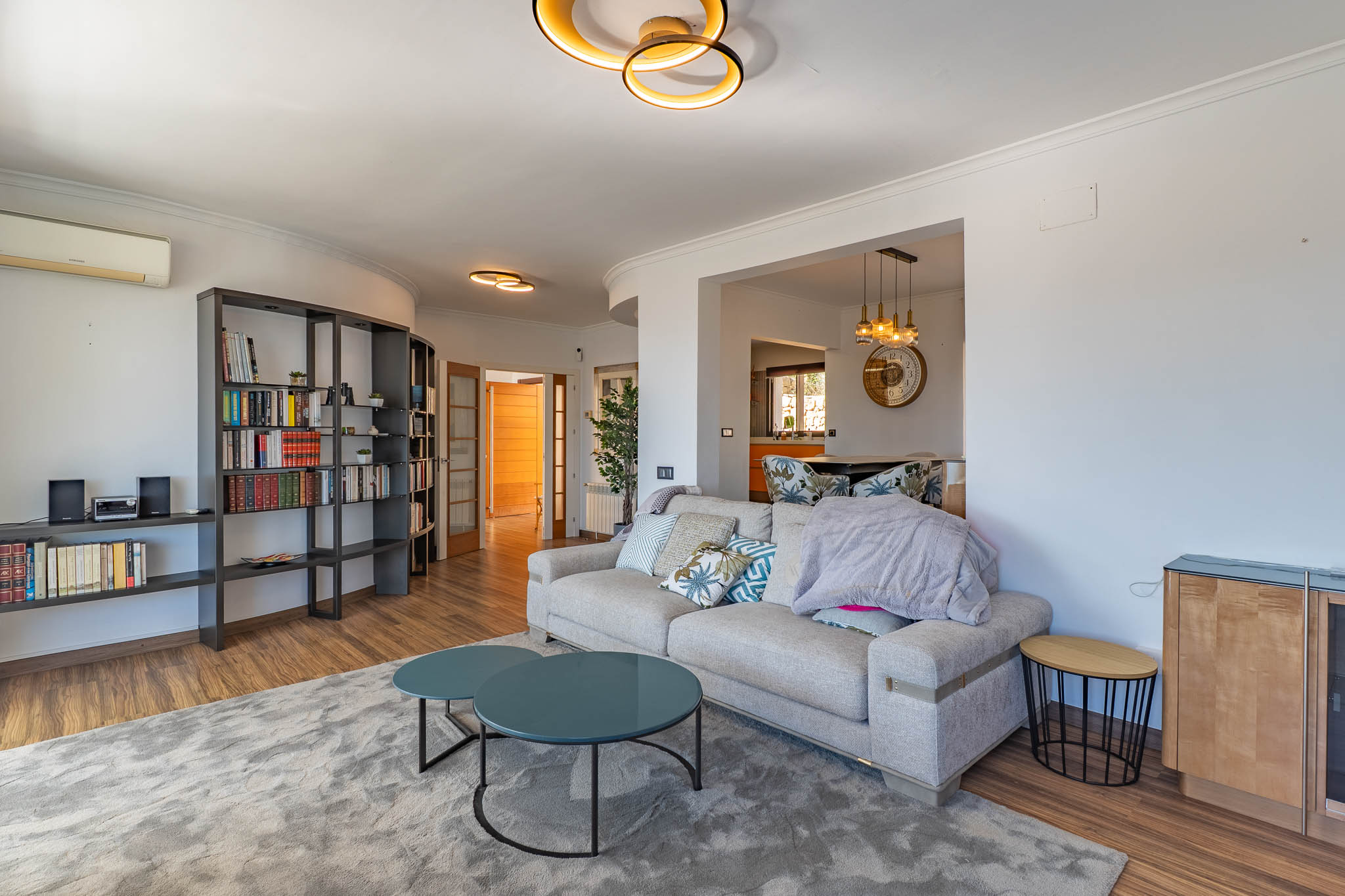 Espacioso salón con sofá contemporáneo, mesas de centro redondas en tono teal y una estantería modular oscura. Destacan la iluminación de diseño y los suelos de madera. Espacioso salón con sofá contemporáneo, mesas de centro redondas en tono teal y una estantería modular oscura. Destacan la iluminación de diseño y los suelos de madera.