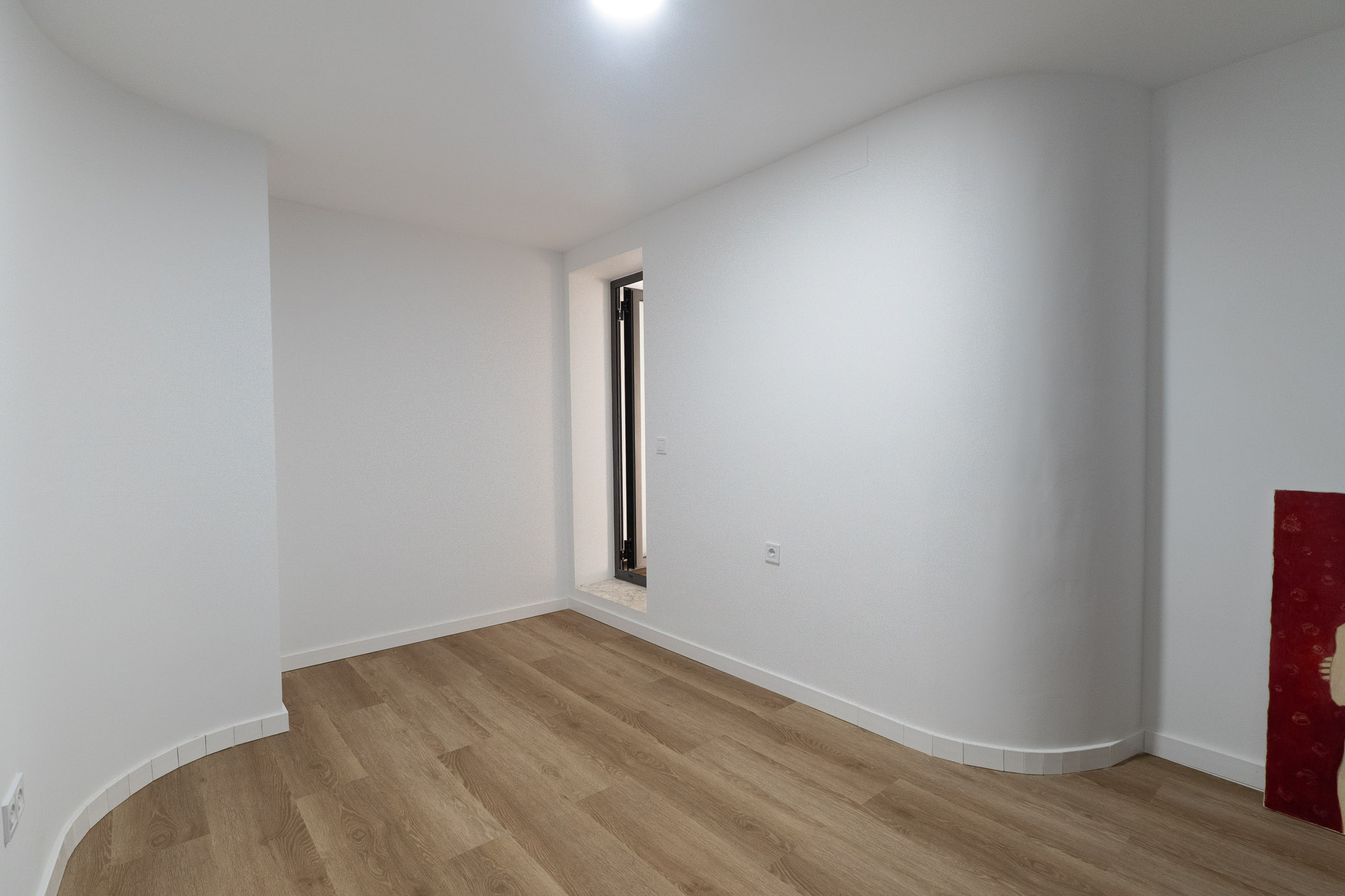Estancia luminosa con suelo de lamas claras, paredes blancas y una singular pared curva. Destaca una abertura vertical con marco oscuro y umbral de piedra. Estancia luminosa con suelo de lamas claras, paredes blancas y una singular pared curva. Destaca una abertura vertical con marco oscuro y umbral de piedra.