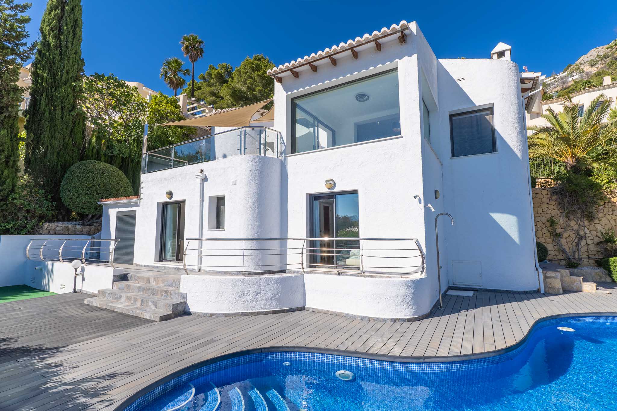 Fachada blanca de la villa con amplias ventanas y terrazas acristaladas. Destaca la piscina de mosaico azul y la tarima de composite gris, enmarcada por cipreses y palmeras. Fachada blanca de la villa con amplias ventanas y terrazas acristaladas. Destaca la piscina de mosaico azul y la tarima de composite gris, enmarcada por cipreses y palmeras.