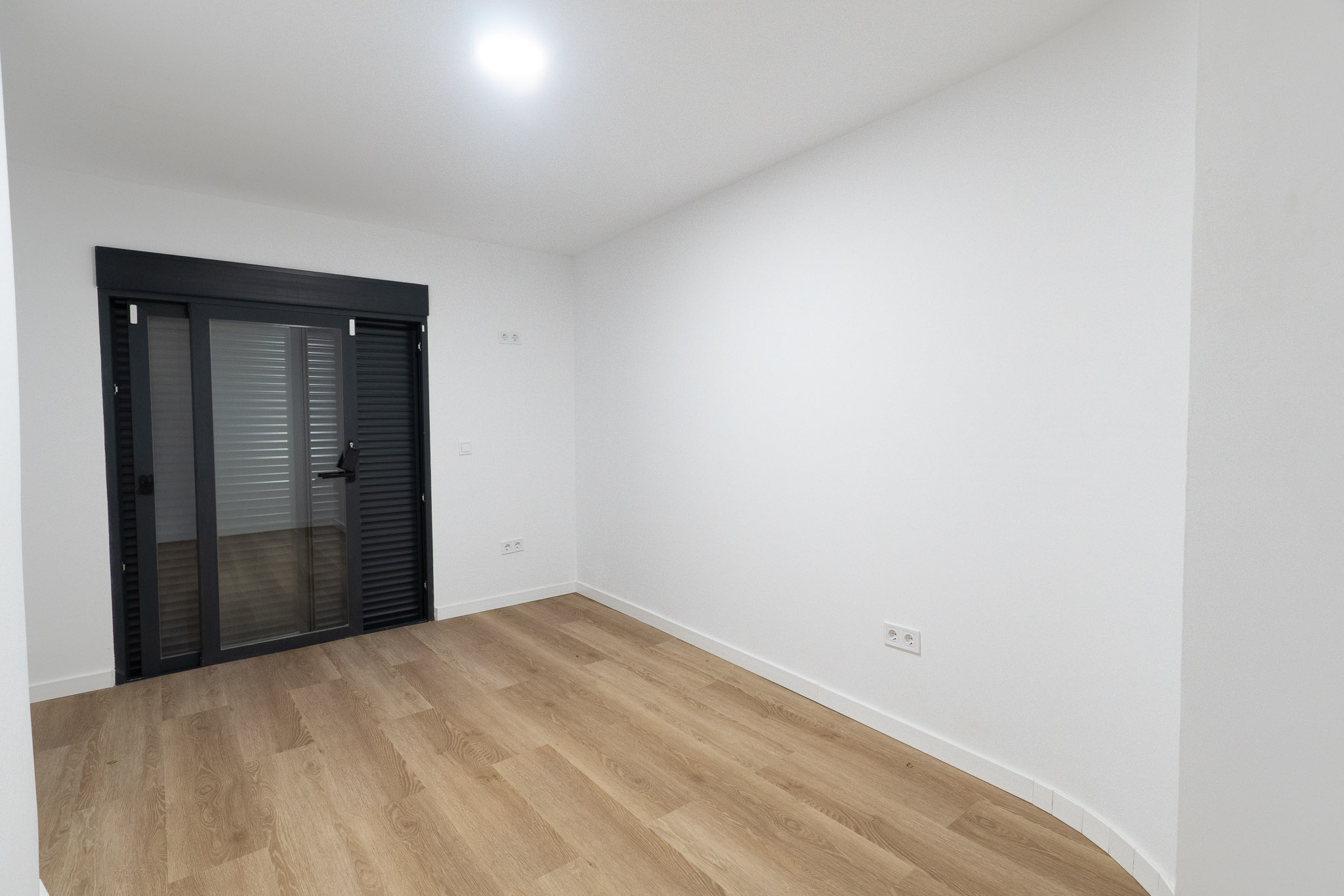 Habitación luminosa con suelo laminado de madera clara y paredes blancas. Destaca una puerta corredera de cristal en gris antracita con persianas integradas que da acceso al exterior. Habitación luminosa con suelo laminado de madera clara y paredes blancas. Destaca una puerta corredera de cristal en gris antracita con persianas integradas que da acceso al exterior.