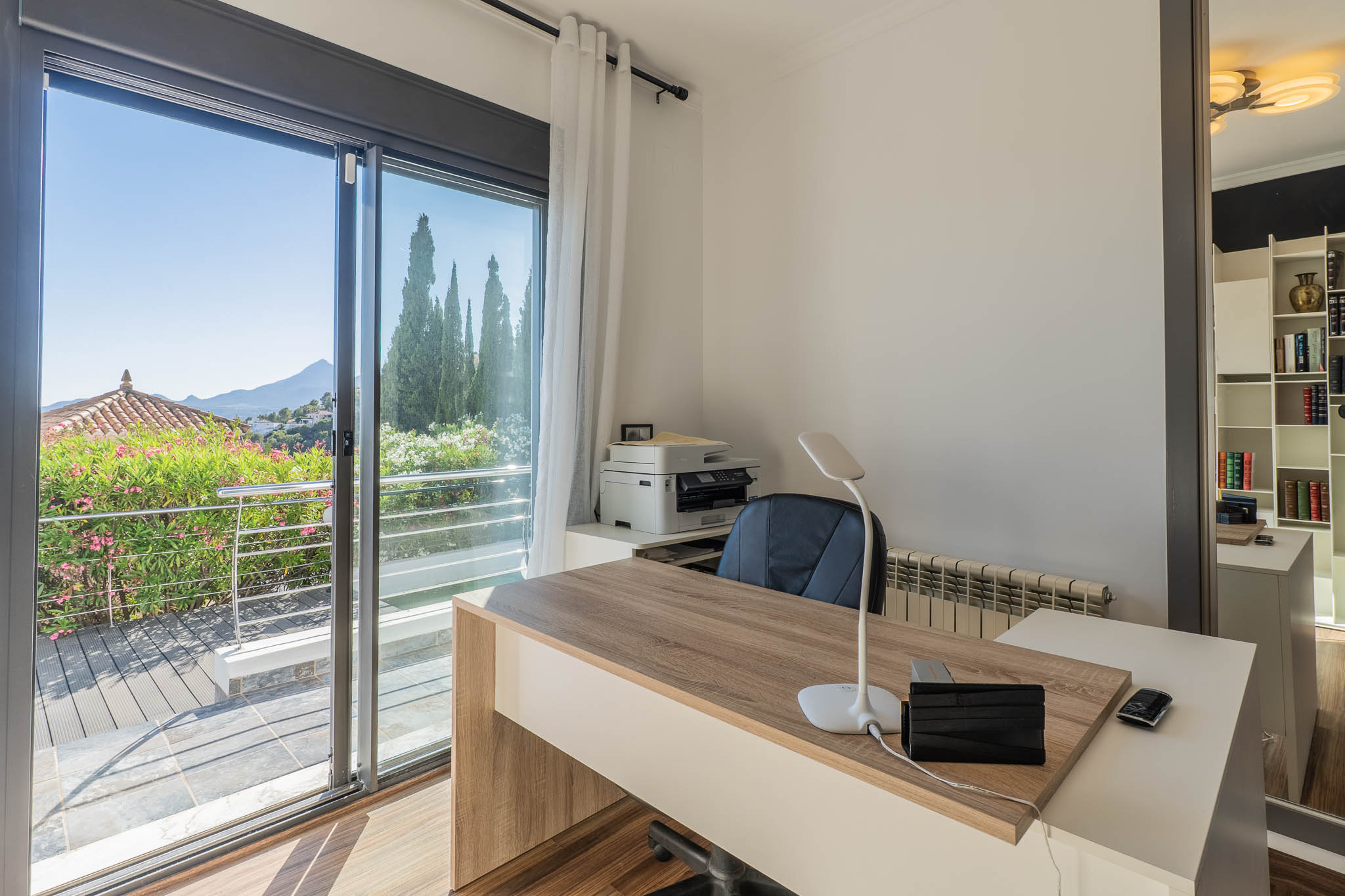 Luminoso despacho con escritorio moderno de madera, gran puerta corredera de cristal a una terraza con vegetación exuberante y vistas panorámicas a la montaña. Un espejo refleja una práctica estantería. Luminoso despacho con escritorio moderno de madera, gran puerta corredera de cristal a una terraza con vegetación exuberante y vistas panorámicas a la montaña. Un espejo refleja una práctica estantería.