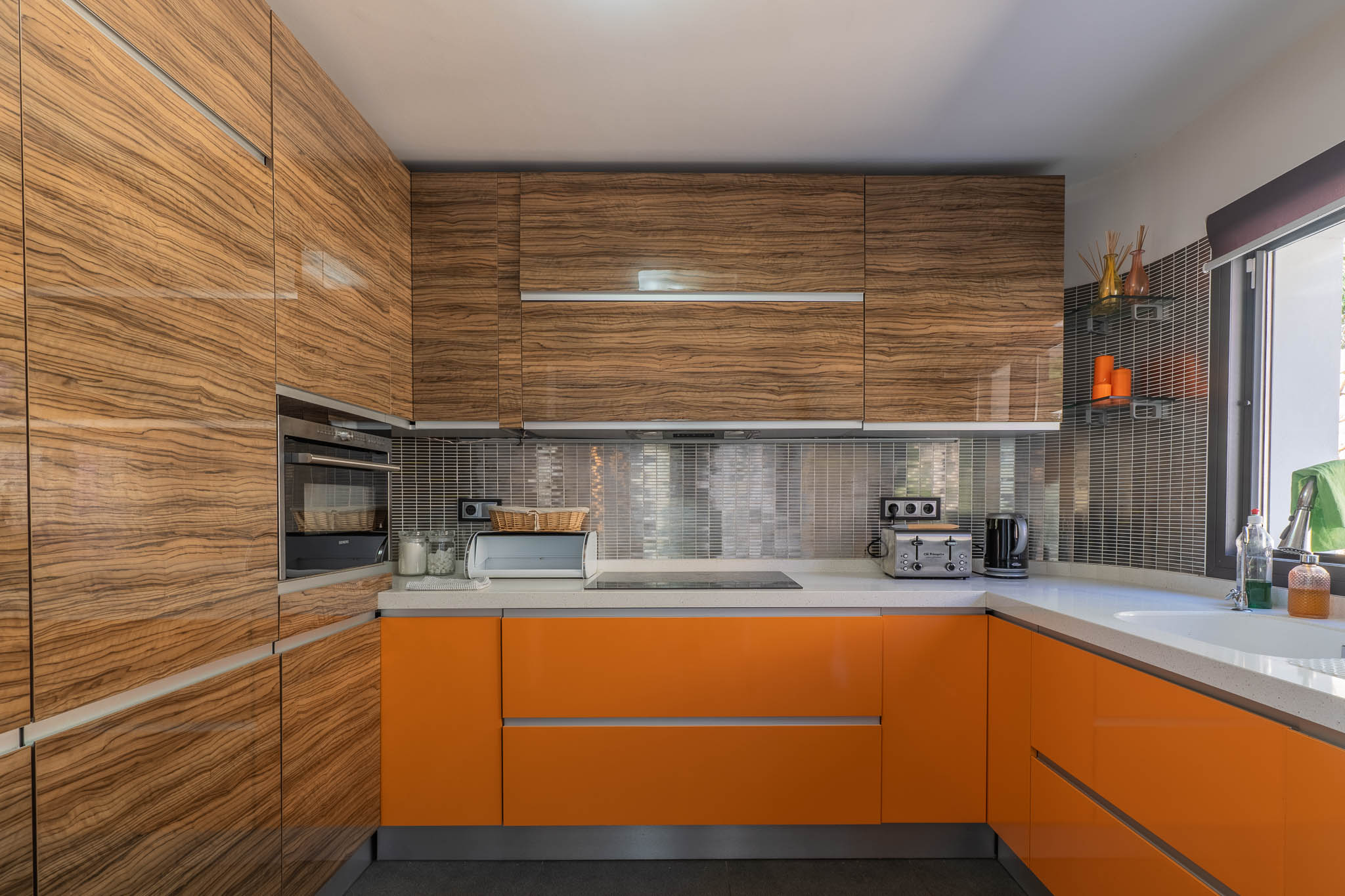 Moderna cocina con armarios superiores de madera veteada brillante y bajos en naranja vibrante, complementada por encimeras claras y un salpicadero de mosaico metálico. Moderna cocina con armarios superiores de madera veteada brillante y bajos en naranja vibrante, complementada por encimeras claras y un salpicadero de mosaico metálico.