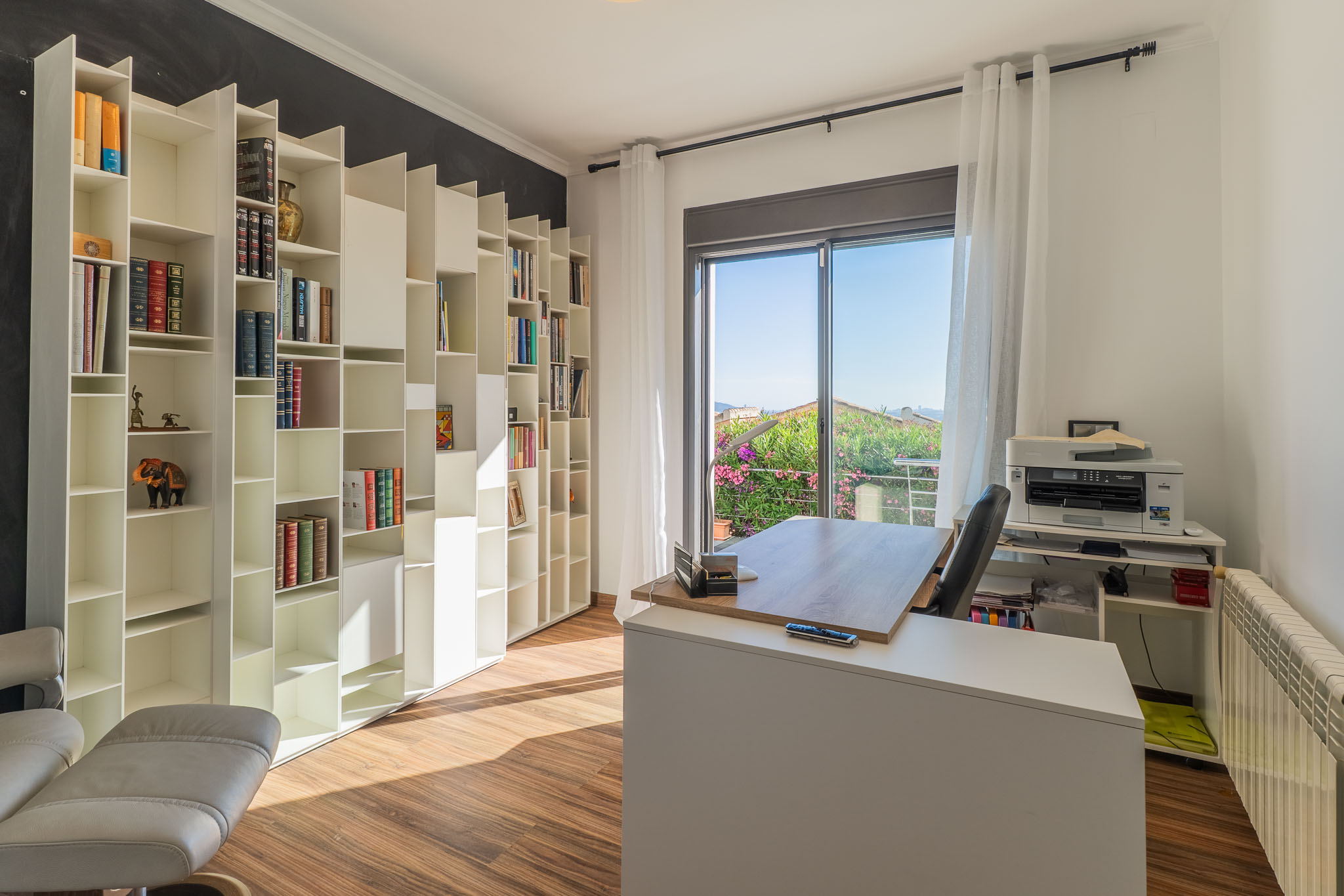 Oficina luminosa con estantería modular blanca, suelo de madera y acceso a balcón con vistas a la vegetación. El escritorio moderno se sitúa frente a la luz natural. Oficina luminosa con estantería modular blanca, suelo de madera y acceso a balcón con vistas a la vegetación. El escritorio moderno se sitúa frente a la luz natural.