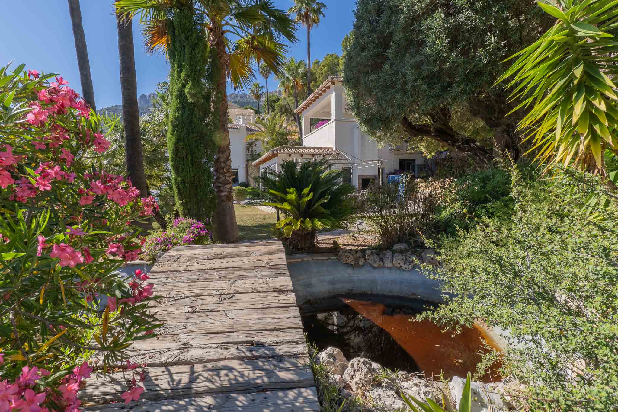 Pasarela de madera rústica sobre un estanque sereno, enmarcada por vibrantes adelfas rosadas y palmeras altas que conducen a la villa bajo el sol mediterráneo. Pasarela de madera rústica sobre un estanque sereno, enmarcada por vibrantes adelfas rosadas y palmeras altas que conducen a la villa bajo el sol mediterráneo.
