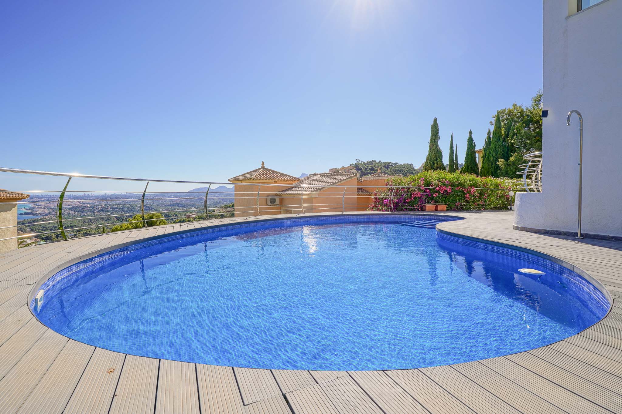 Piscina curva con mosaico azul y tarima de madera clara, enmarcada por una barandilla de acero, ofreciendo vistas panorámicas al valle y ciudad lejana. Piscina curva con mosaico azul y tarima de madera clara, enmarcada por una barandilla de acero, ofreciendo vistas panorámicas al valle y ciudad lejana.