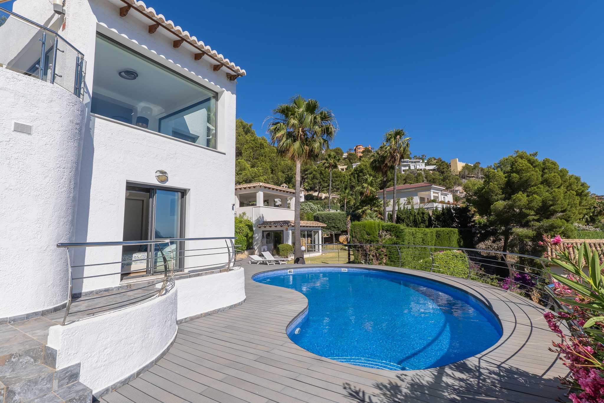 Piscina curva de mosaico azul con tarima de composite gris, adyacente a la fachada blanca con ventanal panorámico y balcón con barandilla de acero. Piscina curva de mosaico azul con tarima de composite gris, adyacente a la fachada blanca con ventanal panorámico y balcón con barandilla de acero.