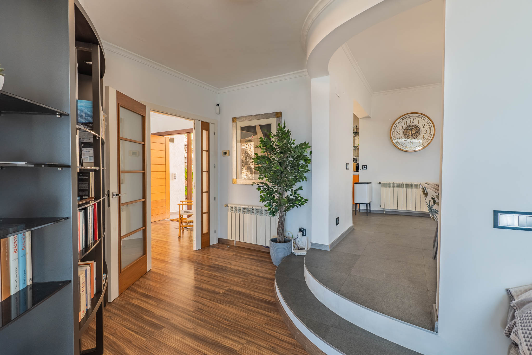 Recibidor con suelo laminado efecto madera que conecta con un espacio exterior a través de puertas acristaladas. Unas escaleras curvas de gres conducen a la zona de comedor, destacando una estantería integrada. Recibidor con suelo laminado efecto madera que conecta con un espacio exterior a través de puertas acristaladas. Unas escaleras curvas de gres conducen a la zona de comedor, destacando una estantería integrada.