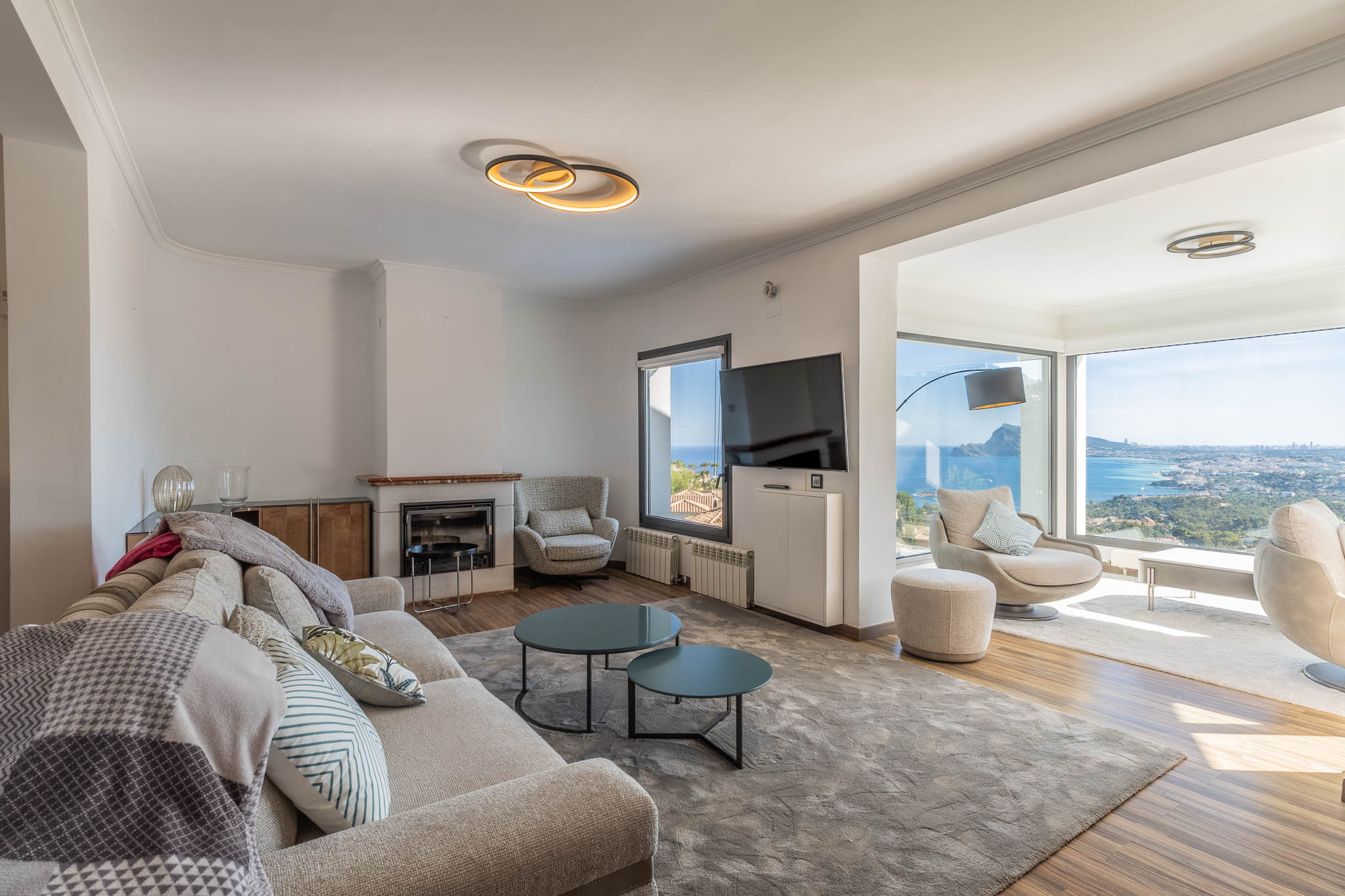 Salón moderno con sofá claro, chimenea y amplios ventanales que ofrecen vistas panorámicas al mar y la ciudad. El mobiliario contemporáneo complementa la luminosidad del espacio. Salón moderno con sofá claro, chimenea y amplios ventanales que ofrecen vistas panorámicas al mar y la ciudad. El mobiliario contemporáneo complementa la luminosidad del espacio.