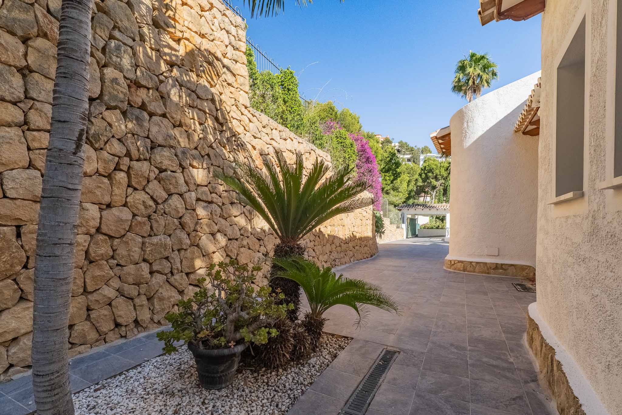 Sendero exterior con muro de piedra natural y vegetación mediterránea, flanqueado por una fachada curva de estuco blanco bajo la luz solar. Sendero exterior con muro de piedra natural y vegetación mediterránea, flanqueado por una fachada curva de estuco blanco bajo la luz solar.