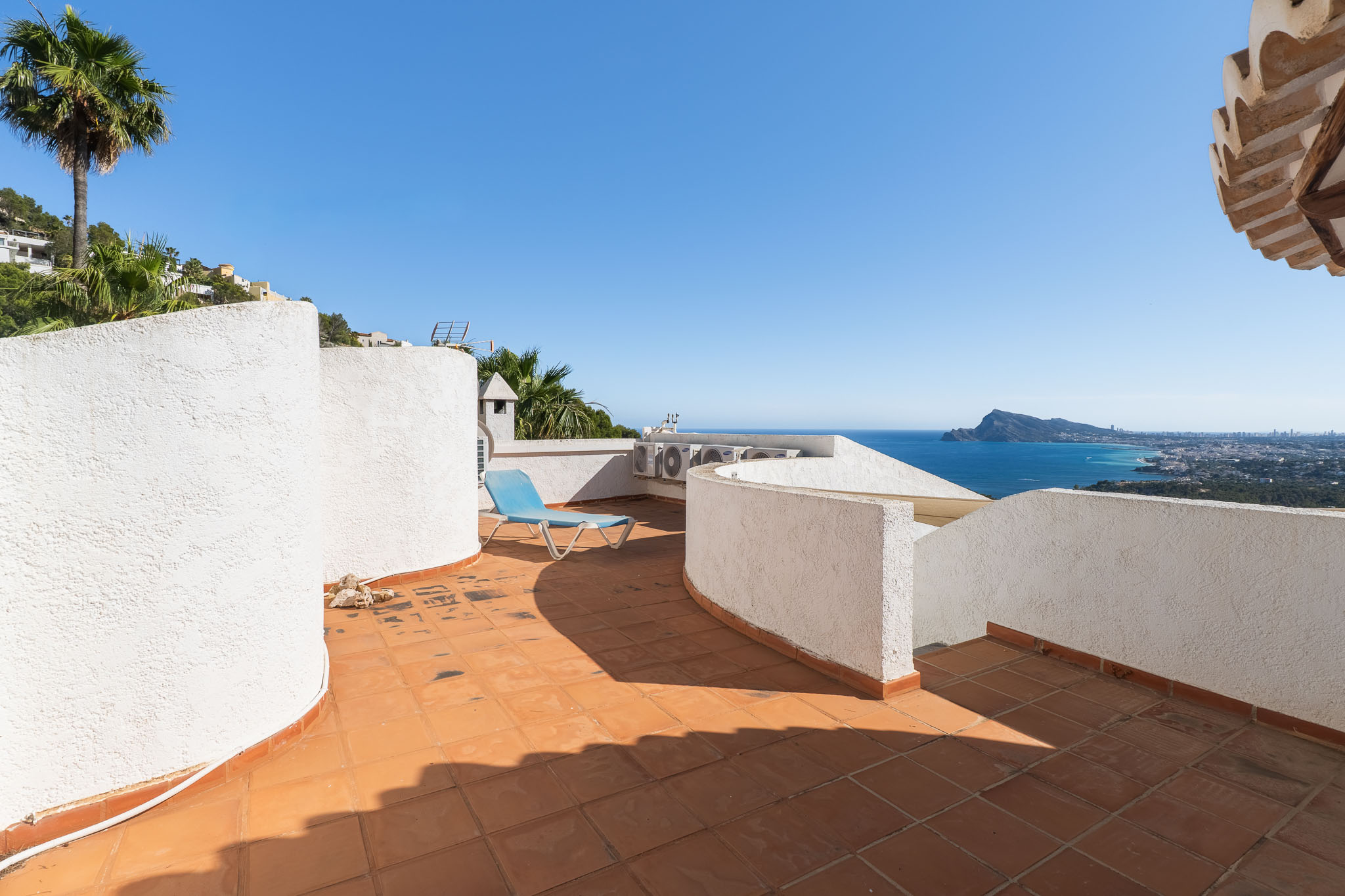 Terraza en la azotea con baldosas de terracota y muros blancos texturizados, ofreciendo vistas panorámicas al mar y la costa. Una tumbona azul invita a la relajación. Terraza en la azotea con baldosas de terracota y muros blancos texturizados, ofreciendo vistas panorámicas al mar y la costa. Una tumbona azul invita a la relajación.