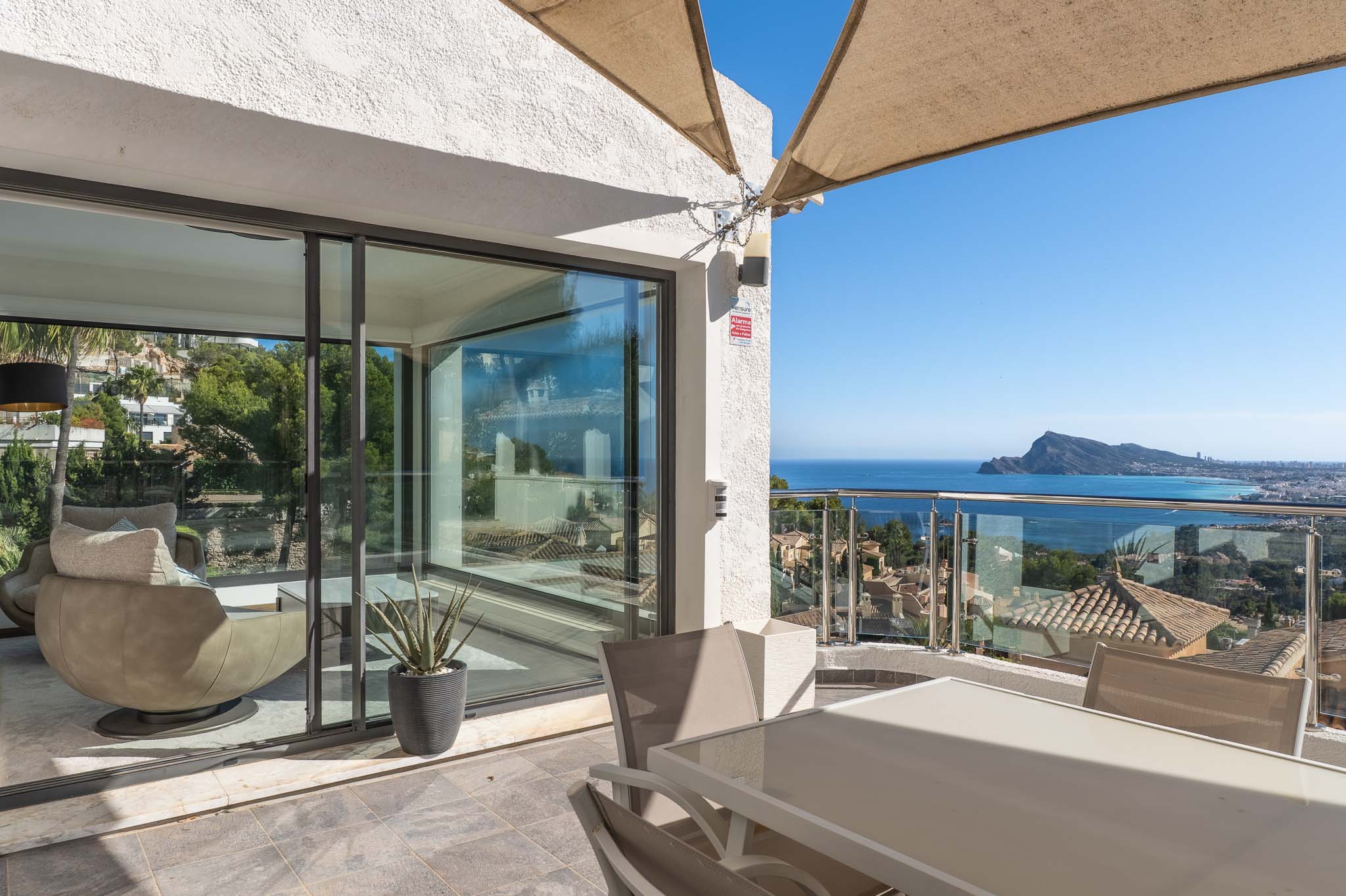 Terraza exterior con mesa de comedor y sillas, protegida por un toldo. Ofrece vistas panorámicas al mar y la costa a través de una barandilla de cristal, conectando con un interior luminoso. Terraza exterior con mesa de comedor y sillas, protegida por un toldo. Ofrece vistas panorámicas al mar y la costa a través de una barandilla de cristal, conectando con un interior luminoso.