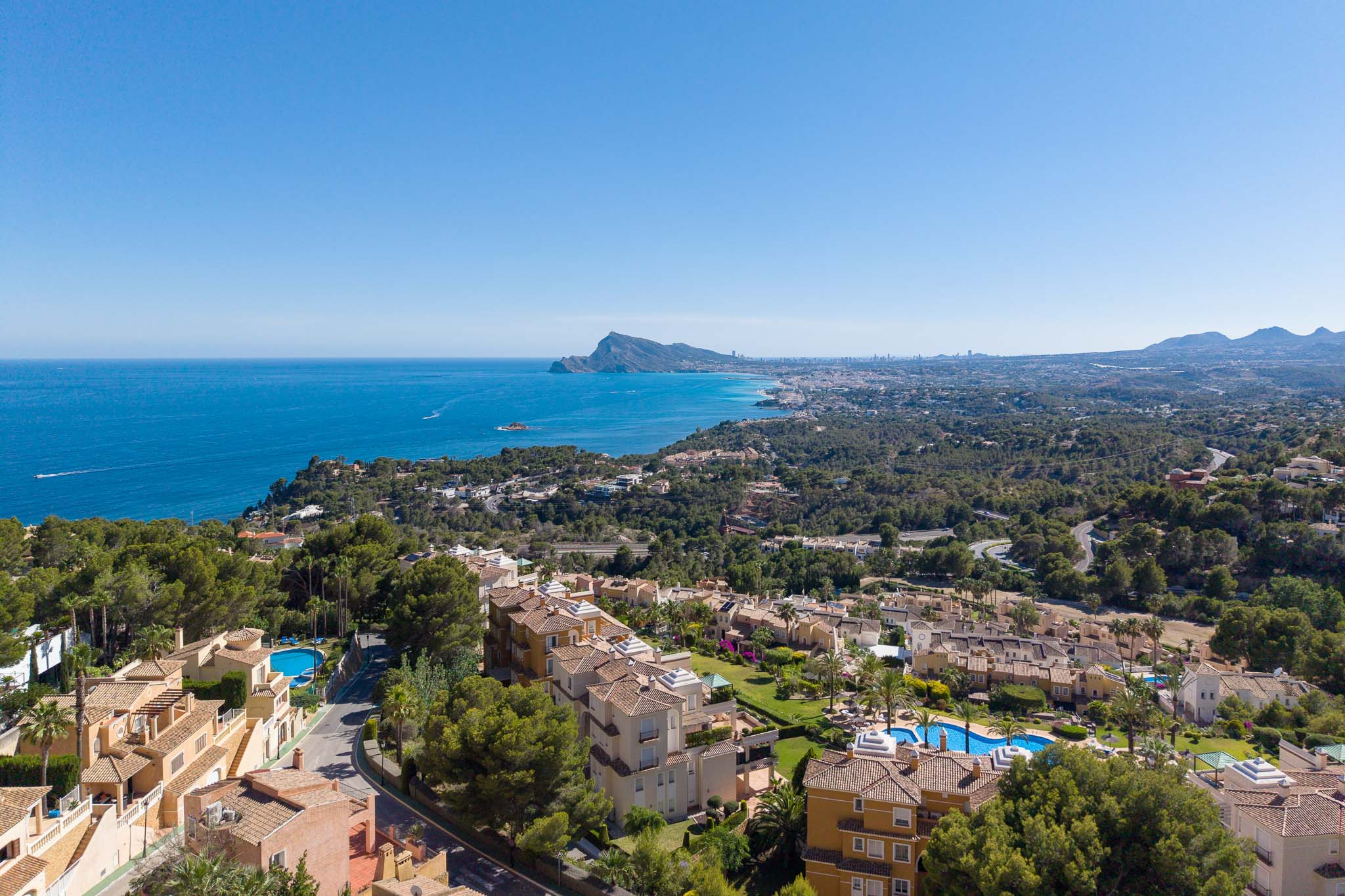 Vista aérea de la costa de Altea Hills, con el mar Mediterráneo, el Peñón de Ifach y la silueta de Benidorm al fondo. Tejados de terracota y piscinas se aprecian entre la vegetación. Vista aérea de la costa de Altea Hills, con el mar Mediterráneo, el Peñón de Ifach y la silueta de Benidorm al fondo. Tejados de terracota y piscinas se aprecian entre la vegetación.