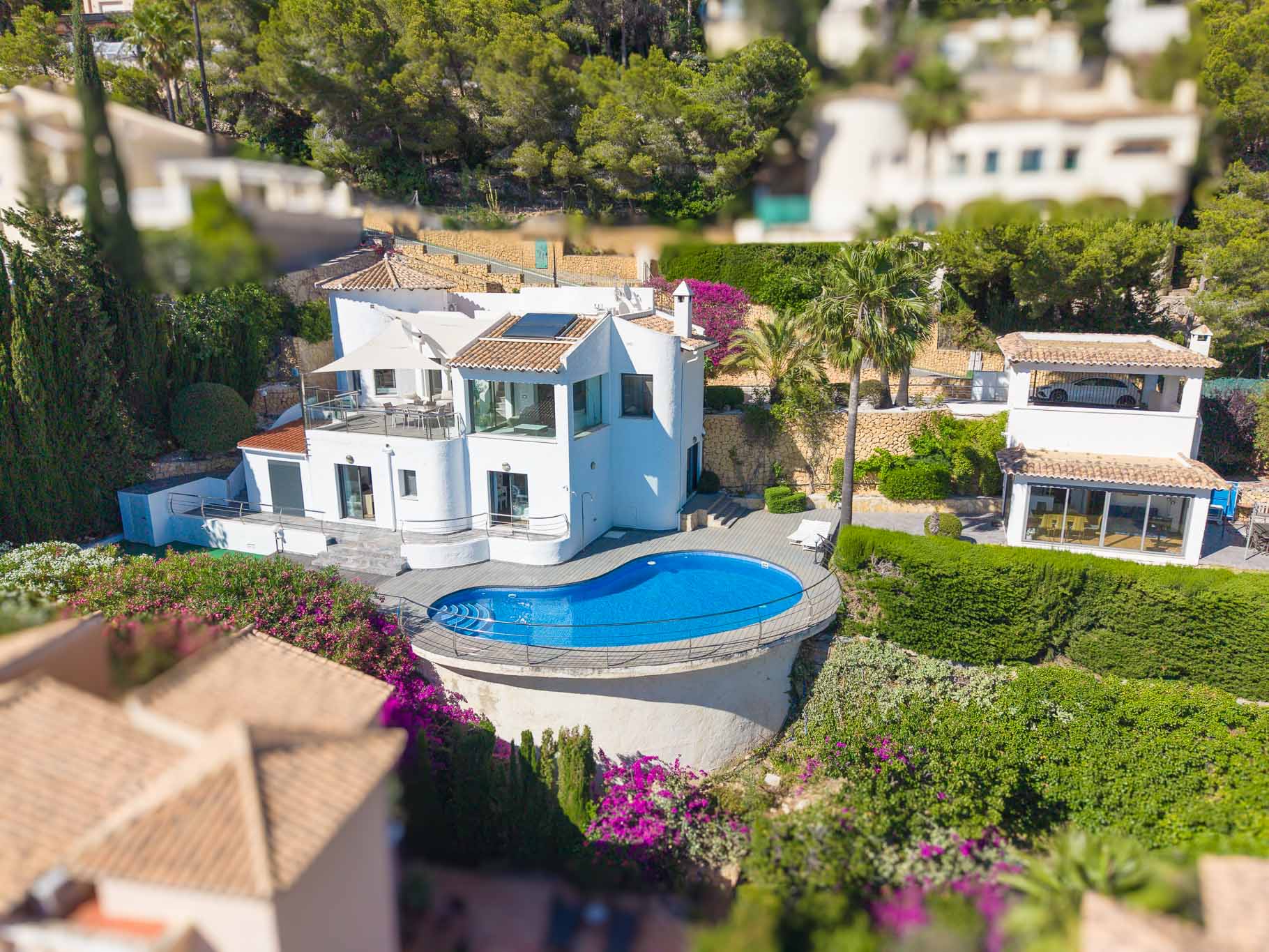 Vista aérea de la villa blanca, destacando la piscina de forma irregular con terraza de madera y barandilla. Muestra múltiples terrazas y el exuberante jardín mediterráneo. Vista aérea de la villa blanca, destacando la piscina de forma irregular con terraza de madera y barandilla. Muestra múltiples terrazas y el exuberante jardín mediterráneo.