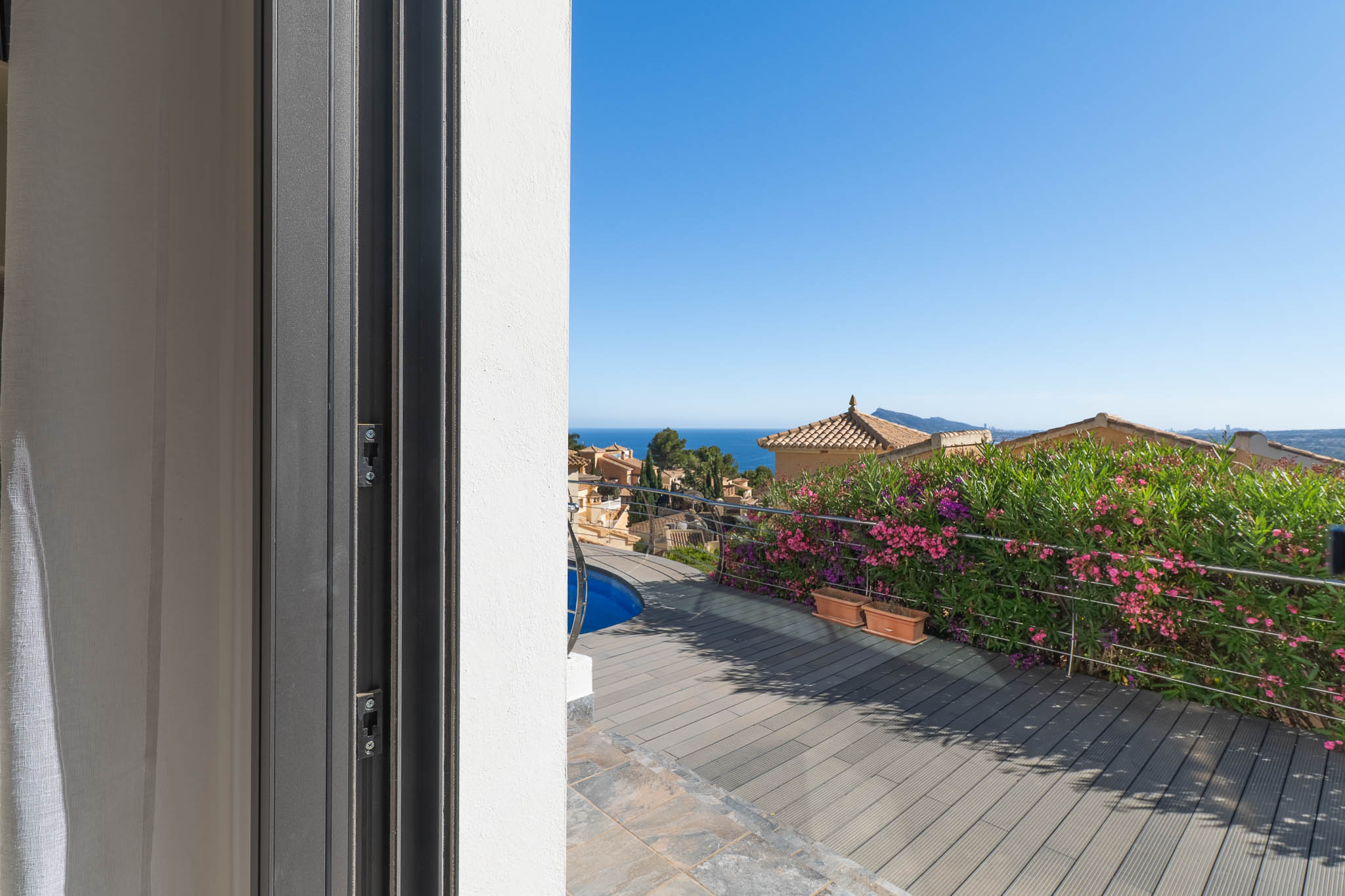 Vista desde la puerta corredera a la terraza con tarima de composite, piscina y barandilla de acero. El mar Mediterráneo y tejados tradicionales se aprecian al fondo. Vista desde la puerta corredera a la terraza con tarima de composite, piscina y barandilla de acero. El mar Mediterráneo y tejados tradicionales se aprecian al fondo.