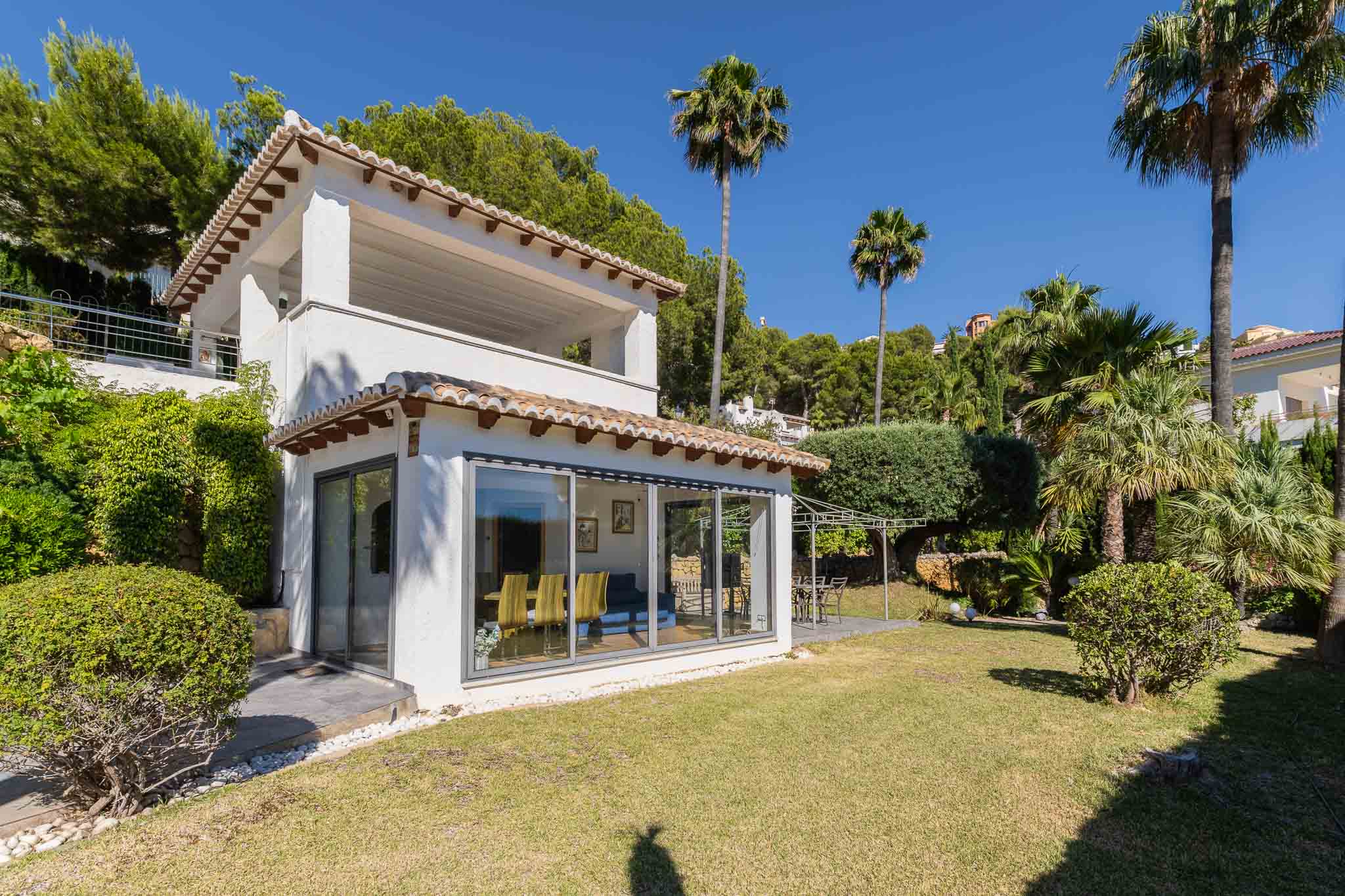 Vista exterior de la villa con fachada blanca, tejas tradicionales y amplios ventanales que conectan el interior con el jardín. Destaca el césped plano y la zona de comedor exterior bajo pérgola. Vista exterior de la villa con fachada blanca, tejas tradicionales y amplios ventanales que conectan el interior con el jardín. Destaca el césped plano y la zona de comedor exterior bajo pérgola.