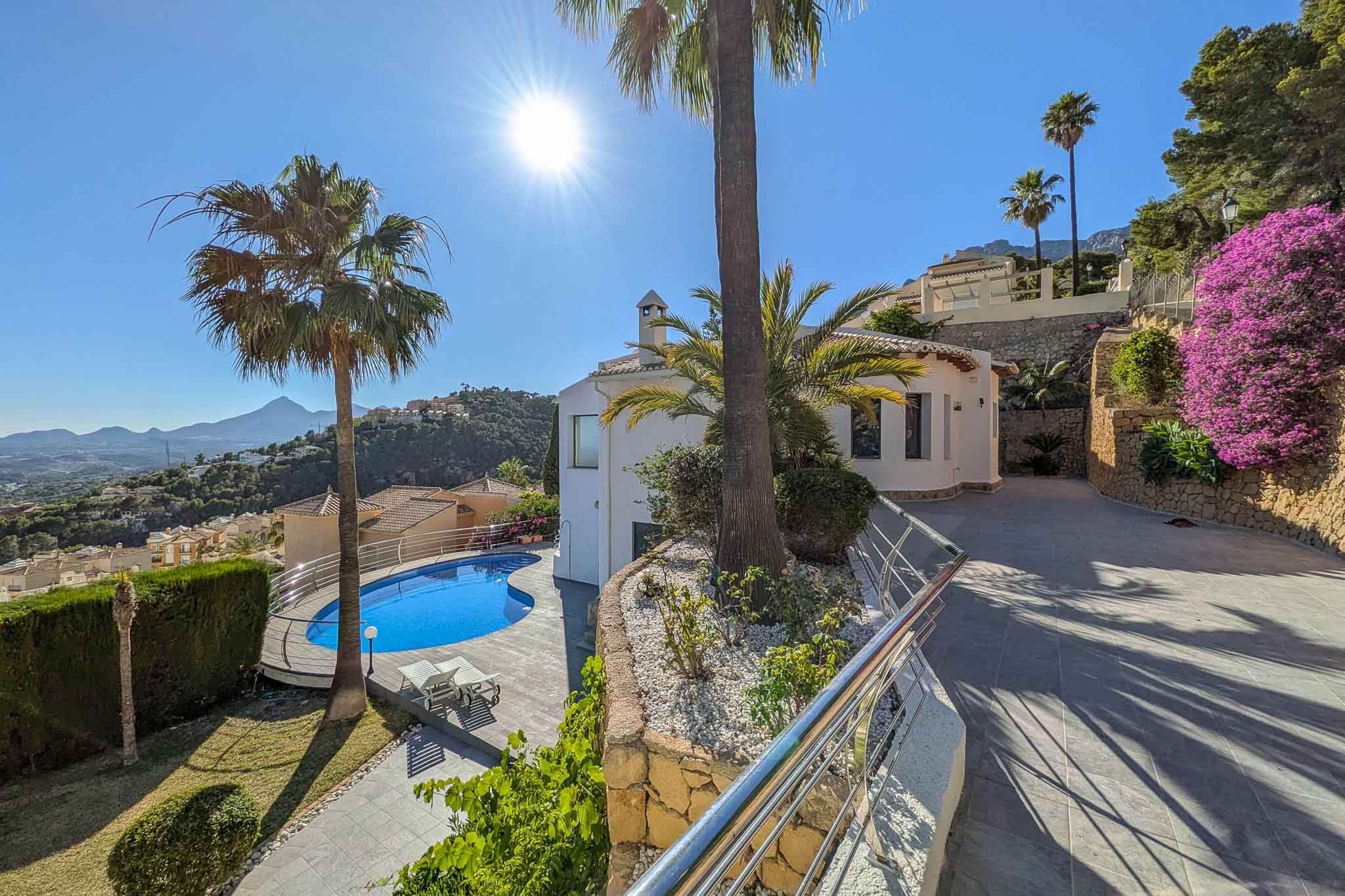 Vista exterior de la villa con piscina de forma irregular, rodeada de palmeras y un cuidado jardín. La entrada pavimentada asciende junto a muros de piedra y buganvillas, ofreciendo vistas a las montañas. Vista exterior de la villa con piscina de forma irregular, rodeada de palmeras y un cuidado jardín. La entrada pavimentada asciende junto a muros de piedra y buganvillas, ofreciendo vistas a las montañas.