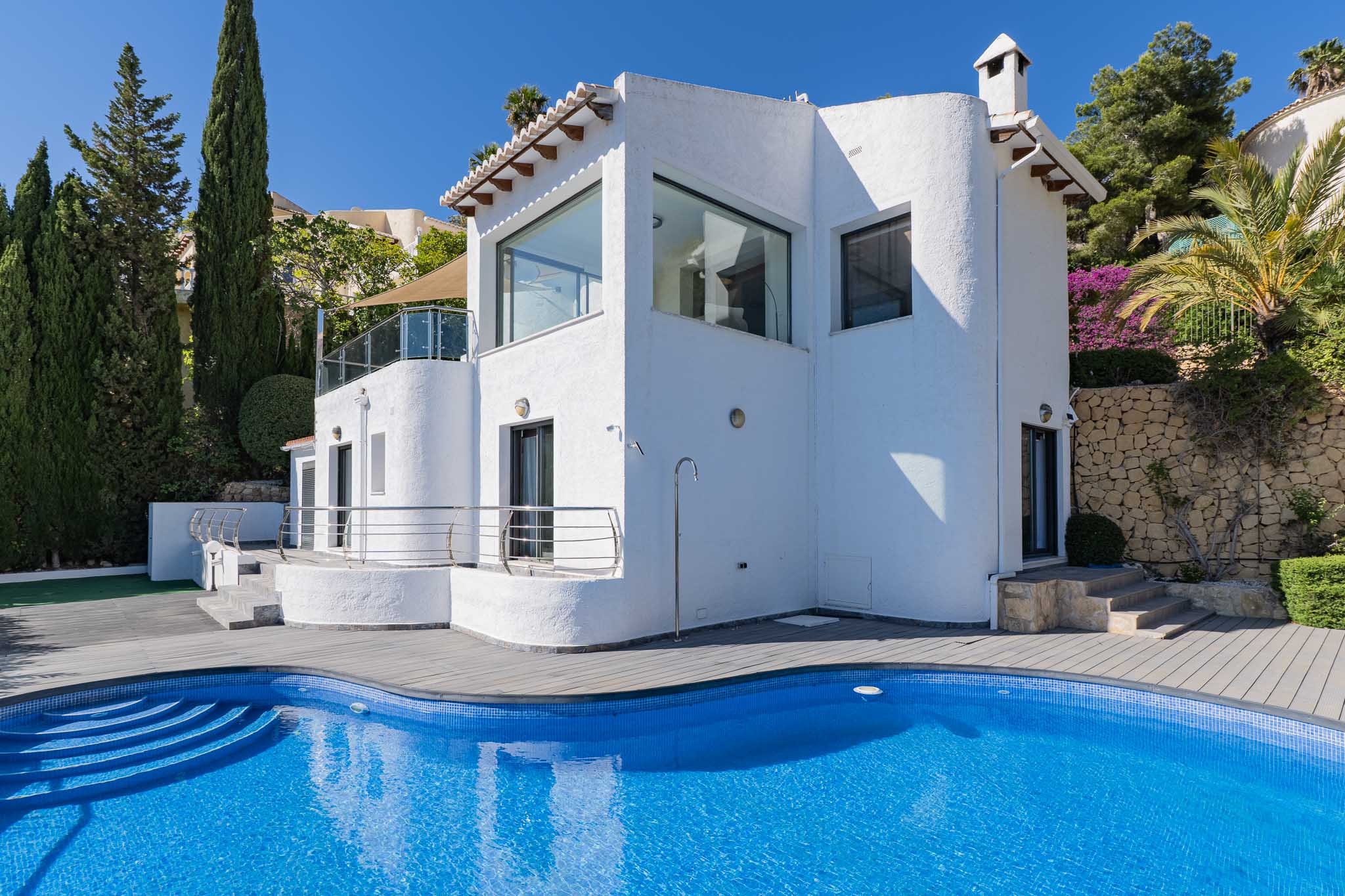 Vista exterior de la villa, destacando una piscina curva de mosaico azul y tarima gris, complementada por una ducha exterior y vegetación mediterránea. Vista exterior de la villa, destacando una piscina curva de mosaico azul y tarima gris, complementada por una ducha exterior y vegetación mediterránea.