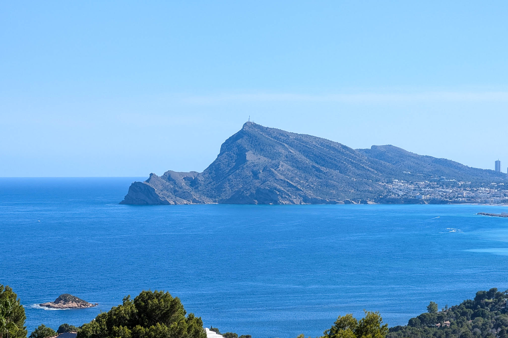 Vista panorámica del Mediterráneo con el imponente perfil de la Sierra Helada, una ciudad costera a lo lejos y pequeños islotes rocosos bajo un cielo azul claro. Vista panorámica del Mediterráneo con el imponente perfil de la Sierra Helada, una ciudad costera a lo lejos y pequeños islotes rocosos bajo un cielo azul claro.