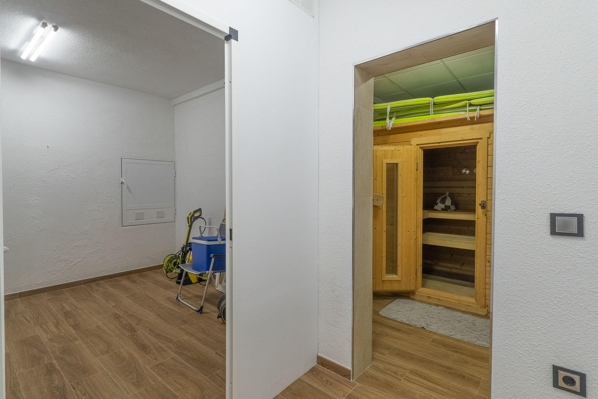 Zona de bienestar con cabina de sauna de madera y bancos interiores, contigua a un cuarto de servicio. Ambos espacios presentan suelo de gres imitación madera. Zona de bienestar con cabina de sauna de madera y bancos interiores, contigua a un cuarto de servicio. Ambos espacios presentan suelo de gres imitación madera.