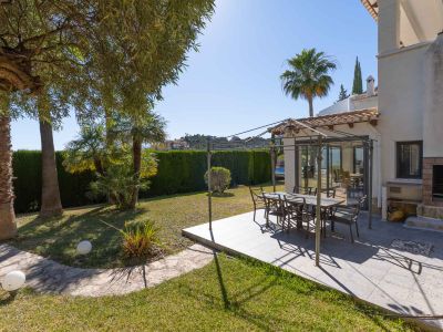 Acogedor comedor exterior con pérgola metálica sobre patio embaldosado, ideal para comidas al aire libre. Se complementa con una barbacoa de obra y un frondoso jardín con palmeras y seto de privacidad.