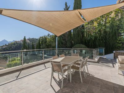 Amplia terraza exterior con vela de sombra, equipada con mesa de comedor y barandilla de cristal, ofreciendo vistas panorámicas a las montañas y la urbanización.