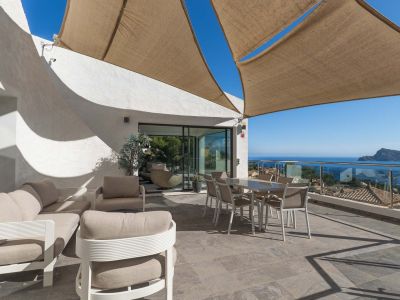 Amplia terraza exterior con zona de estar y comedor bajo vela de sombra, ofreciendo vistas panorámicas al mar y la costa a través de barandilla de cristal.