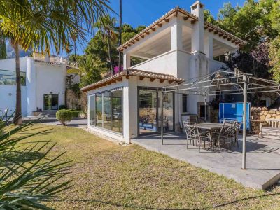 Amplia terraza pavimentada con comedor exterior bajo pérgola metálica, adyacente a una sala acristalada y rodeada de un cuidado jardín mediterráneo.
