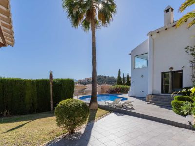Amplio jardín con piscina circular y solárium de tarima, flanqueado por una villa blanca y un gran seto que ofrece privacidad. Vistas a las colinas.