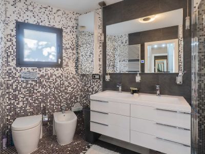 Baño con paredes y suelo de mosaico en tonos tierra y gris. Destacan el doble lavabo blanco con mueble suspendido, un gran espejo iluminado y sanitarios murales, complementados por un radiador toallero.