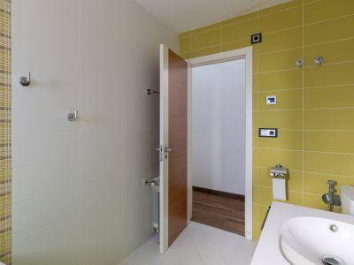 Baño de cortesía con diseño moderno, destacando azulejos en tonos beige y verde lima, un lavabo contemporáneo y detalles en cromo. La puerta de madera entreabre a un espacio con suelo de parquet.