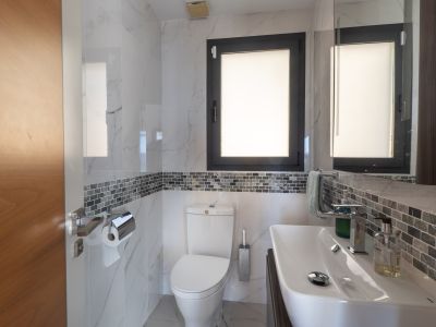 Baño moderno con azulejos efecto mármol blanco, una banda decorativa de mosaico gris y ventana con marco oscuro. Incluye lavabo blanco y sanitarios a juego.