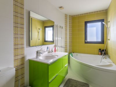 Baño moderno con bañera de esquina y lavabo verde brillante. Azulejos blancos texturizados y verdes lima, con un detalle de mosaico a cuadros. Ventana con marco oscuro y toallero calefactable.