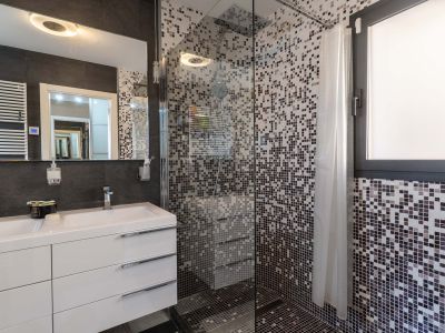 Baño moderno con doble lavabo, mueble blanco y espejo iluminado. La ducha de cristal presenta mosaico en tonos grises y una ventana integrada.