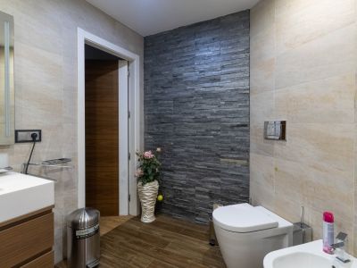 Baño moderno con pared decorativa de piedra apilada oscura, azulejos beige claro y suelo de imitación madera. Incluye inodoro, bidé y tocador con lavabo.