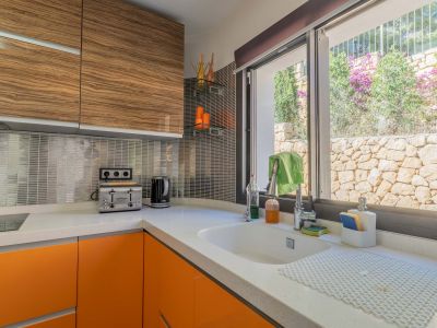 Cocina moderna con encimera de cuarzo blanco, armarios bajos naranjas y superiores de madera veteada. Ventana con vistas a un muro de piedra natural y vegetación.
