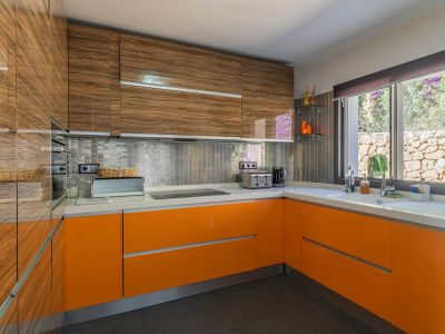Cocina moderna con mobiliario superior de madera veteada y bajos lacados en naranja brillante. Destacan la encimera blanca, el salpicadero de mosaico metálico y la ventana con vistas a un muro de piedra.
