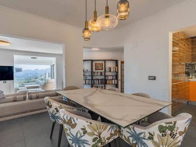 Comedor de diseño con mesa de efecto mármol y sillas tapizadas, iluminado por lámparas colgantes modernas. Conecta con el salón, ofreciendo vistas panorámicas a la montaña.