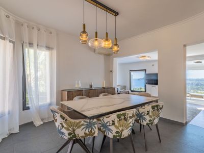 Comedor moderno con mesa de mármol y sillas de diseño tropical. Un candelabro lineal ilumina el espacio, que se abre a vistas panorámicas al mar desde las estancias contiguas.