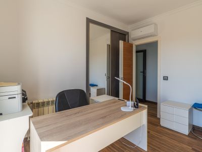 Despacho luminoso con escritorio de madera clara, suelo laminado y armario con puertas correderas de espejo, equipado con aire acondicionado.