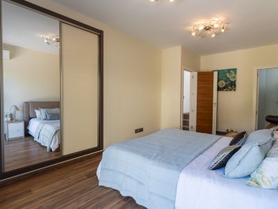 Dormitorio con cama de textiles azules, suelo laminado de madera y armario corredero con panel espejado. Destaca la moderna lámpara de techo y un cuadro floral.