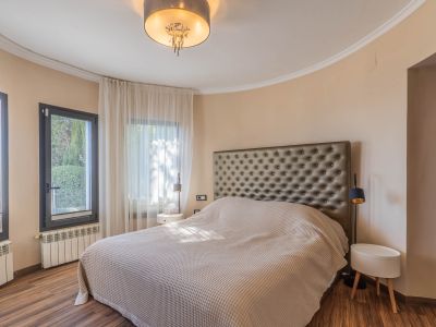 Dormitorio con cama grande y cabecero tapizado. Pared curva con ventanales de marco oscuro y cortinas claras. Suelo de madera, radiadores y lámpara de techo moderna.