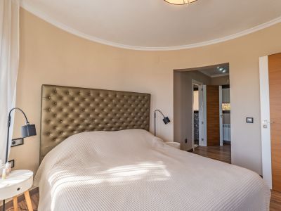 Dormitorio principal con cabecero tapizado, pared curva y luz natural filtrada por cortinas translúcidas, complementado con puertas de madera.