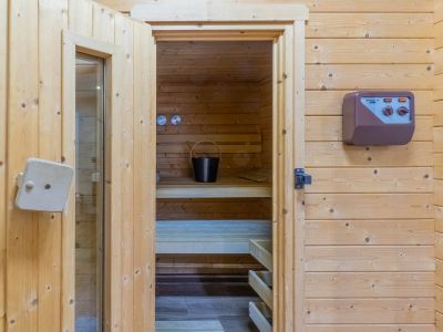 Entrada a la sauna revestida en madera clara, mostrando bancos interiores, un cubo con cucharón y un panel de control externo.